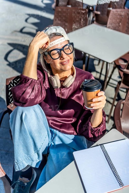 Stylish woman relaxing at an outdoor cafe with eyes closed, savoring hot coffee from a disposable cup, headphones around her neck and an open notebook on the table