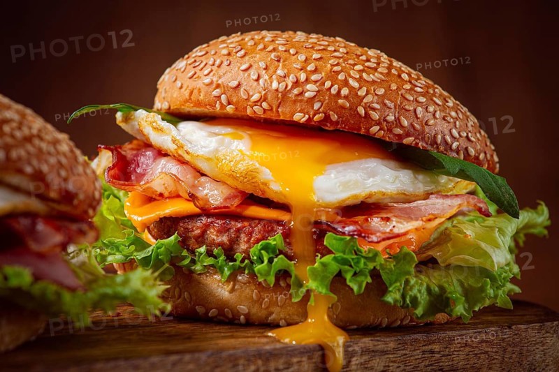 Two juicy burgers sit on a wooden board, each filled with lettuce, bacon, cheese, and fried eggs