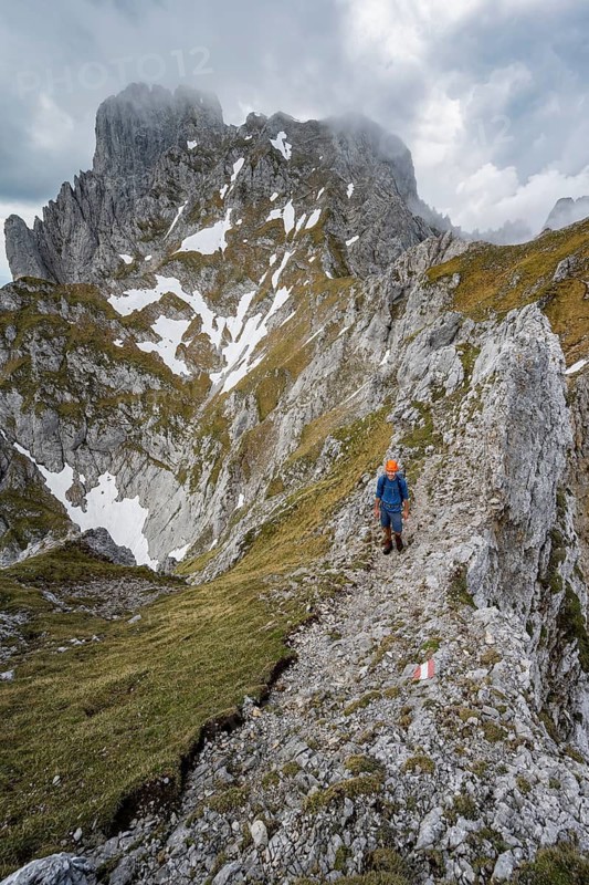 Mountaineer with helmet on a narrow hiking trail on a rocky ridge, ascent to the Maukspitze, clouds moving around the mountains, Ackerlpitze in the background, Wilder Kaiser, Kaiser Mountains, Tyrol, Austria
