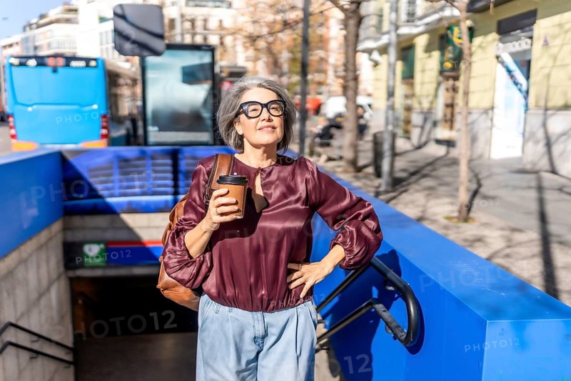 Senior woman with gray hair and glasses stands at a subway entrance holding coffee, looking up at the sun over the city after her morning commute, relaxed and confident