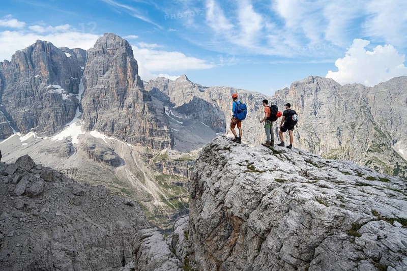 Three mountaineers on a rock in front of a picturesque mountain landscape with rocky peaks, Via Ferrata SOSAT via ferrata, summit of Cima Tosa in the back, Brenta Mountains, Trentino, Italy