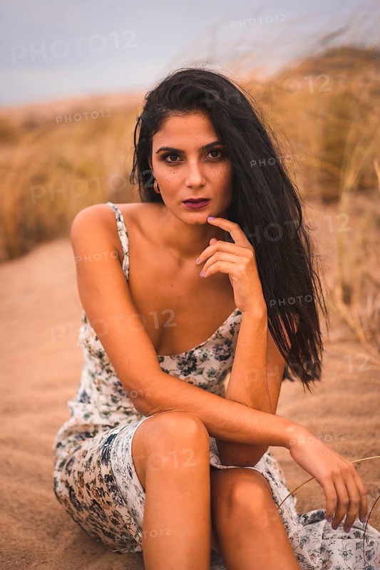 Portrait of a brunette Caucasian caressing her hair in a white floral dress, sitting on the sand on the beach of Cabo de Gata, Nijar. Andalucia, Spain