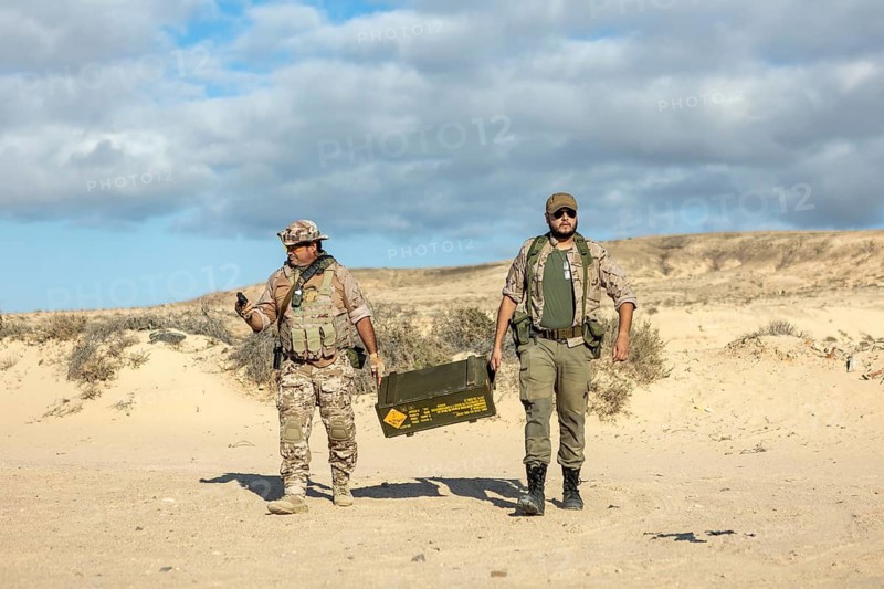 Two soldiers in camouflaged uniforms transport equipment during a military training exercise in a desert environment