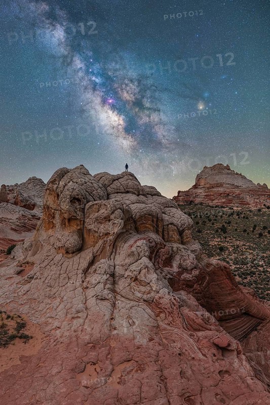 A distant unrecognizable hiker stands atop unique rock formations in Utah, USA, beneath a dazzling Milky Way sky