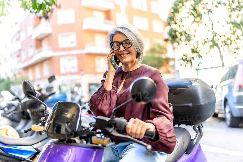 Mature woman on a scooter in a bustling city street, smiling and talking on her smartphone while enjoying an active, independent urban lifestyle and daytime commute