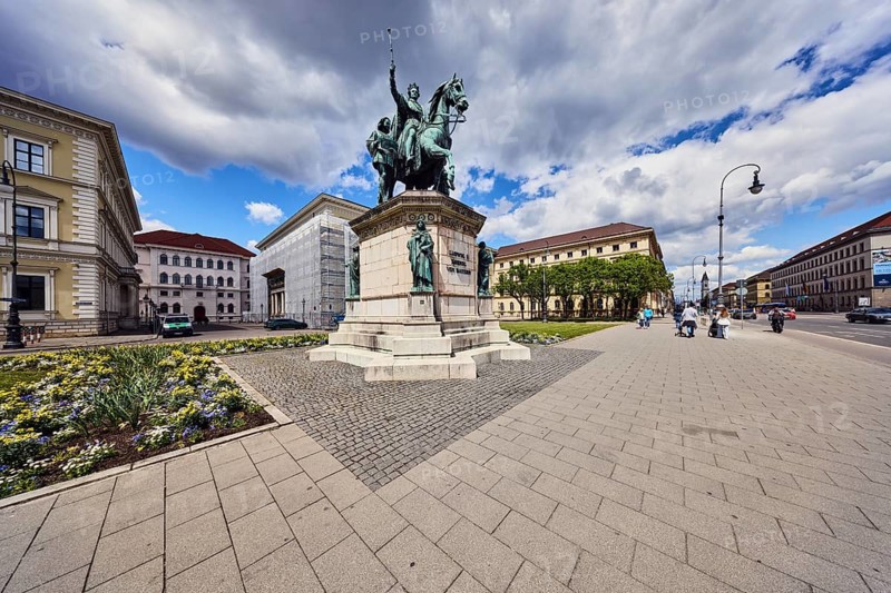Equestrian statue of Ludwig I of Bavaria, sculptor Max Carl von Widnmann, neurainessance style, general architecture, historic buildings, lantern, sidewalk made of pavement tiles, flower bed, trees, pedestrians as a secondary motif, blue sky, white and dark gray cumulus clouds, Odeon Square, paving stones, Munich, state capital, district-free city, Bavaria, Germany