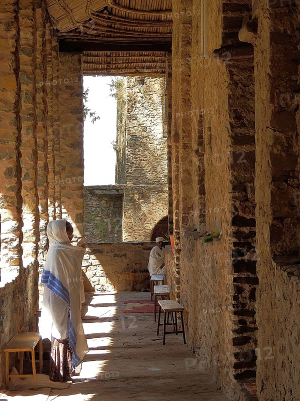 People wearing traditional clothing stand in a shady stone corridor, Debre Birhan Selassie Church, Fasil Ghebbi, Historic Stone Building, Orthodox Church with People Gathering Under Sunny Sky, Gondar, Ethiopia