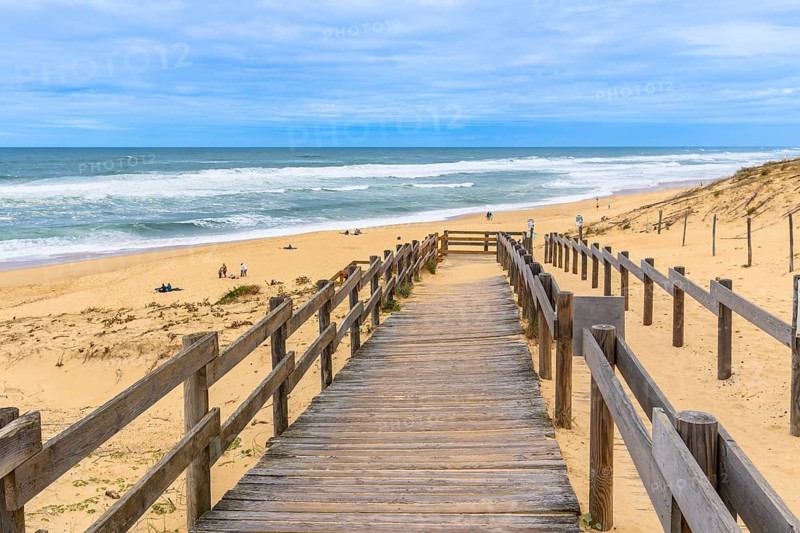 Wooden boardwalk path leading down to the sandy messanges beach, with people enjoying the ocean waves and golden sand along the atlantic coast in landes, nouvelle aquitaine, france
