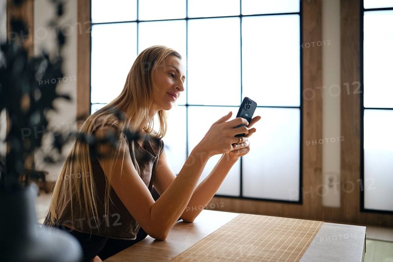 A woman sits at a wooden table in a room with large windows. She is focused on her phone, smiling softly. The room has natural light coming through the windows, creating a bright atmosphere