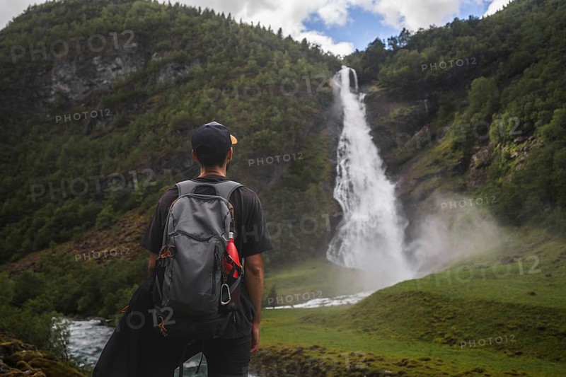 A captivating scene of a traveler with a backpack looking at a cascading waterfall amidst lush green mountains Ideal for themes of exploration, adventure, and nature in Norway