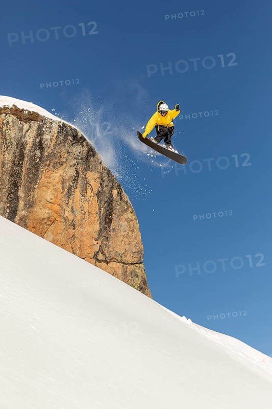 A snowboarder in a yellow jacket executes a thrilling jump off a cliff, sending snow flying against a clear blue sky. Experience the exhilaration of backcountry freeride