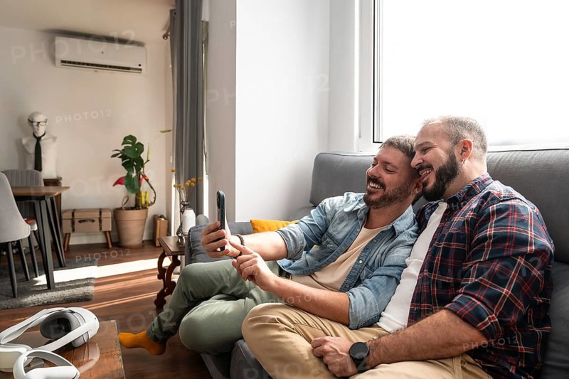 A joyful gay couple takes a selfie on a cozy couch in their modern living room, which features natural light and contemporary decor, creating a warm, intimate atmosphere