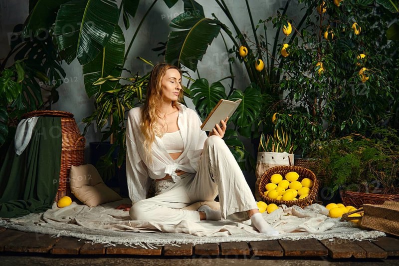 A woman sits on the floor with a book in hand. She is surrounded by green plants and lemon trees. She is relaxed and deeply engaged in reading
