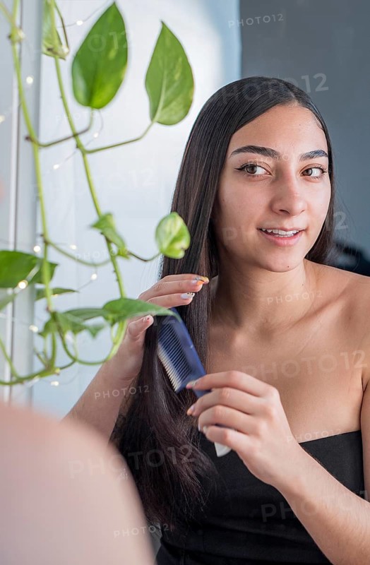 A woman with long, straight hair stands near a window, using a blue comb. She is smiling naturally, surrounded by vibrant green leaves, emphasizing calm and self care