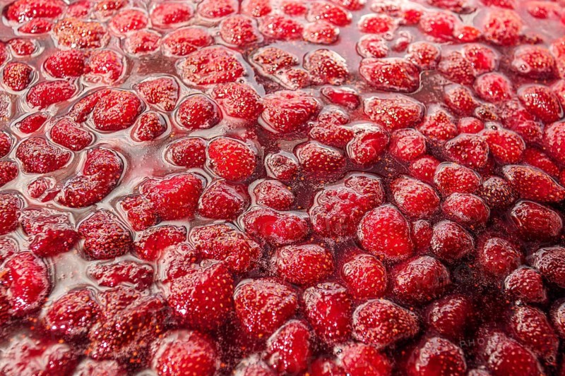 A close up shot of homemade strawberry jam, showcasing its vibrant red color and glistening texture. The jam is filled with whole strawberries, reflecting a delightful sweetness