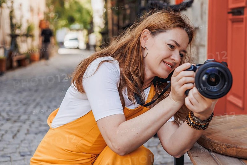 Woman traveler joyfully explores the historic streets of an old town in Turkey, capturing memories with her camera. She exudes happiness, dressed in a white top and orange overalls