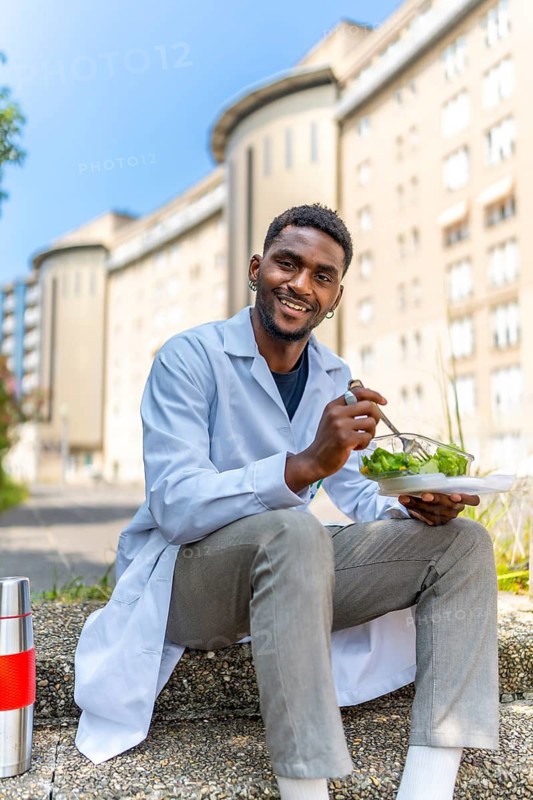 Vertical photo of a young african doctor eating healthy salad on a break outside the hospital