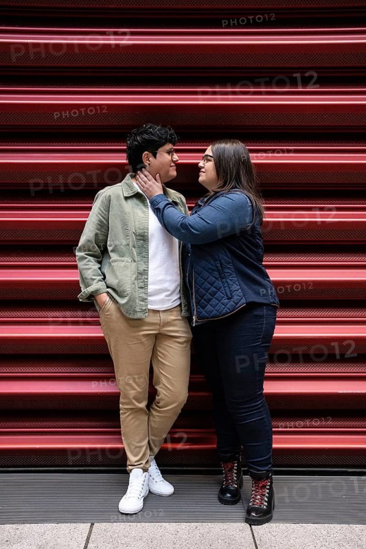 A loving curvy lesbian couple stands close, sharing an intimate moment against a vibrant red backdrop in an urban environment, capturing love and diversity