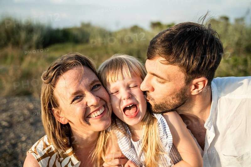 A joyful family, with a mother, father, and daughter, sharing a moment of happiness on an Italian beach. The father gives a kiss as they all smile brightly