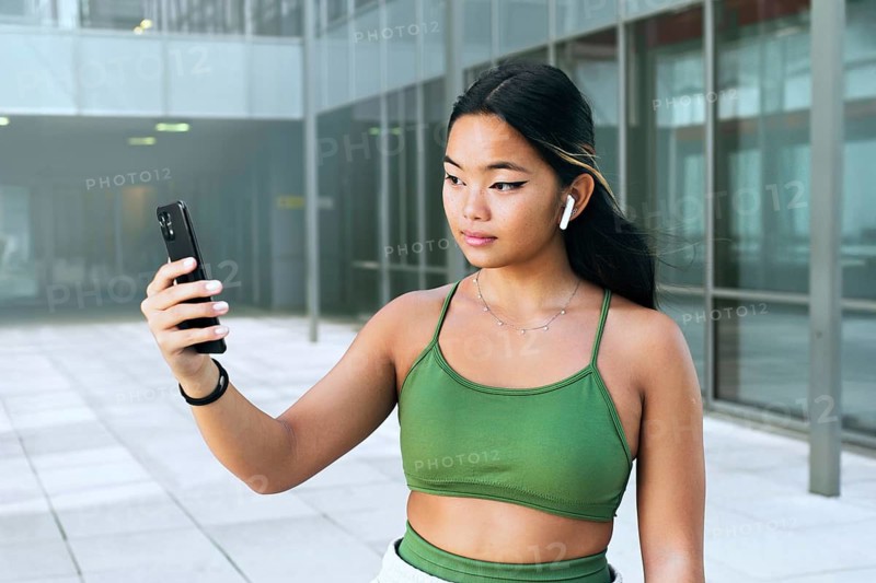 A young Chinese teenager holds a smartphone, wearing wireless earbuds. She is outside an urban building, exuding confidence and modernity in a green top