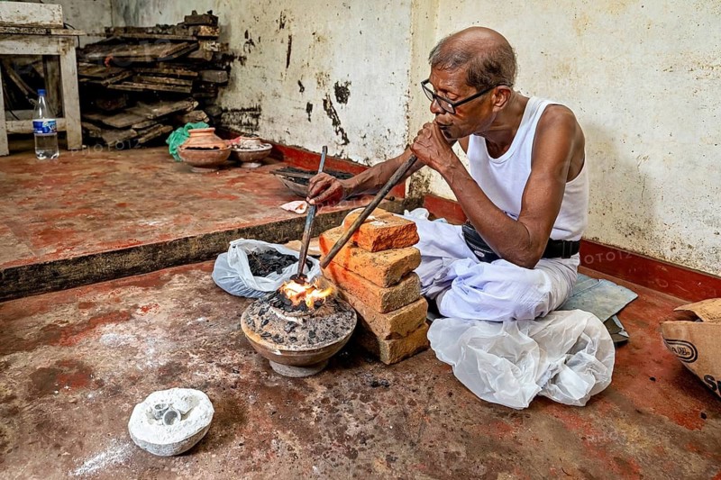 Man blows a flame with a pipe using a traditional workshop technique, a man burns sapphires in a small oven in the town of Ratnapura in Sri Lanka