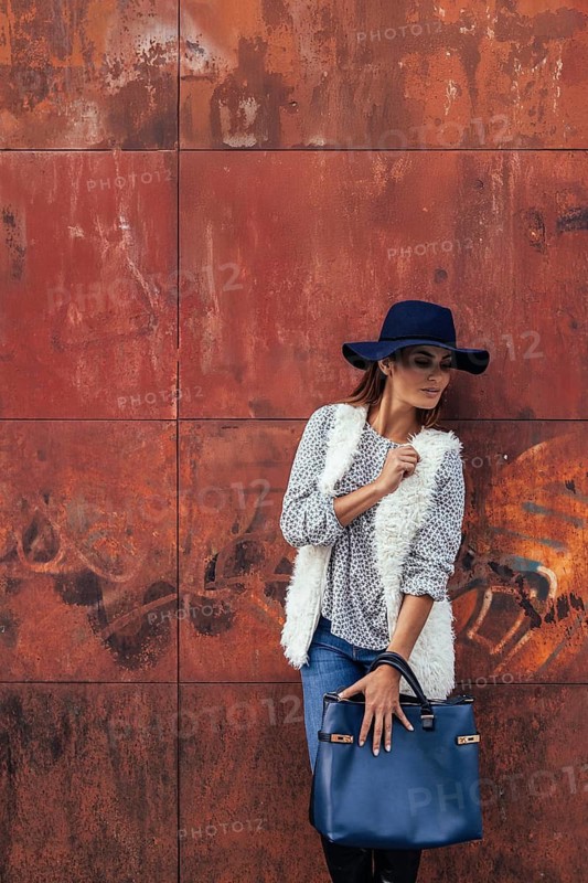 A fashionable woman stands in front of a textured rusted wall, wearing a floppy hat, a fluffy vest, and holding a blue handbag. The scene exudes casual elegance and style