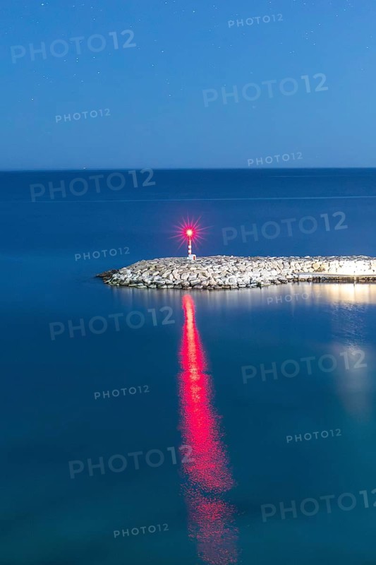 A peaceful scene of the Mediterranean Sea at dusk featuring a solitary lighthouse on a rocky pier. The calm waters reflect the light, creating a serene coastal landscape