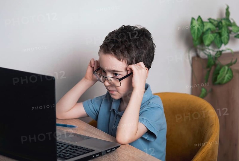 A young child in a blue shirt focuses intently on a laptop screen while adjusting glasses, seated at a wooden table. Indoor setting with a cozy chair and a plant in the background