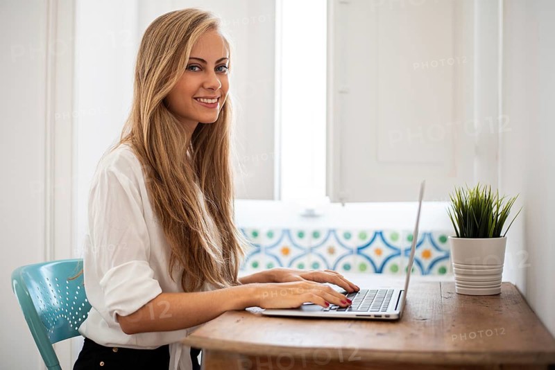 Woman with long blonde hair sits at a wooden desk using a laptop. The scene is bright, with natural light coming through the window, and a small plant beside her