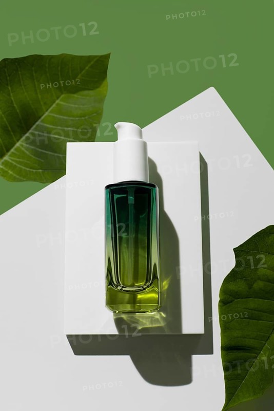 A green glass bottle of essential oil rests on a modern white stand against a striking green background, flanked by vibrant leaves, emphasizing the product's natural appeal for skin care