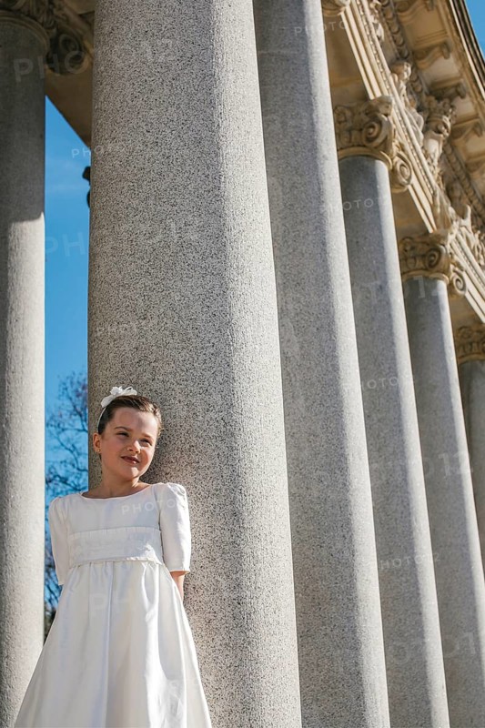 A young girl wearing a First Communion dress stands elegantly beside large stone columns, basking in sunlight. The scene exudes purity and innocence, highlighting a special moment