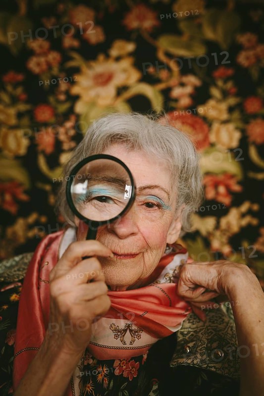 An elderly woman holds a magnifying glass to her eye, set against a vibrant floral backdrop. Her vintage clothing and friendly expression create an inviting, warm ambiance