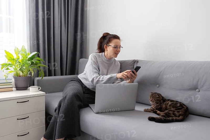 A woman works from home on a grey couch, using her laptop and smartphone. A cat relaxes beside her, and a potted plant and coffee cup are nearby. The setting is cozy and calm