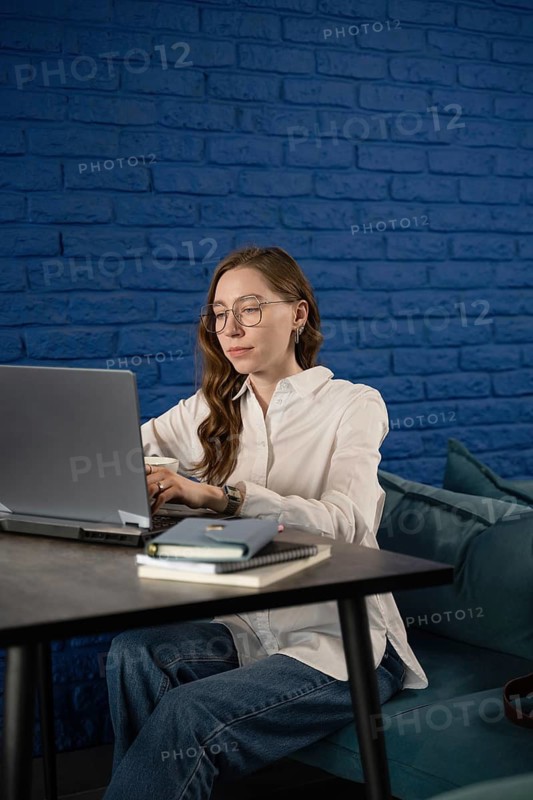 A woman in a white shirt and glasses focuses intently on her laptop in a stylish cafe with a blue brick wall. Notebooks and cushions add a touch of comfort