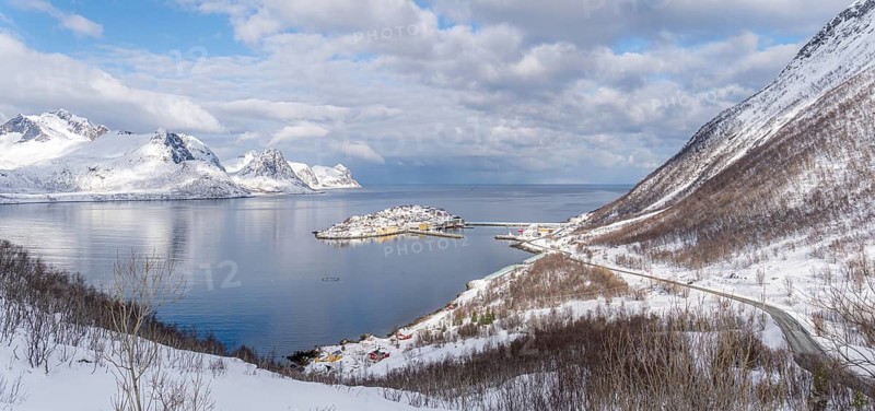 A stunning winter landscape of Husoy, Norway, featuring snow-capped mountains, a serene island village, and the tranquil blue waters of the fjord under a partly cloudy sky