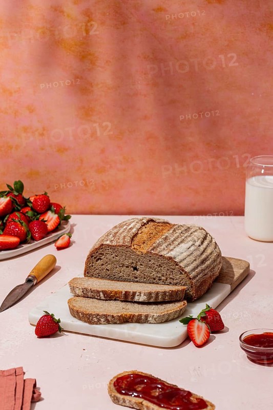 A rustic loaf of bread is sliced on a cutting board, surrounded by fresh strawberries and a pot of jam on a pink table, evoking a summer brunch vibe