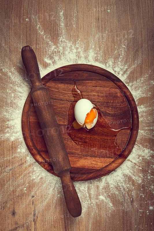 A cracked egg sits on a wooden board with a rolling pin beside it, flour scattered around, capturing a moment of cooking preparation in a cozy kitchen.