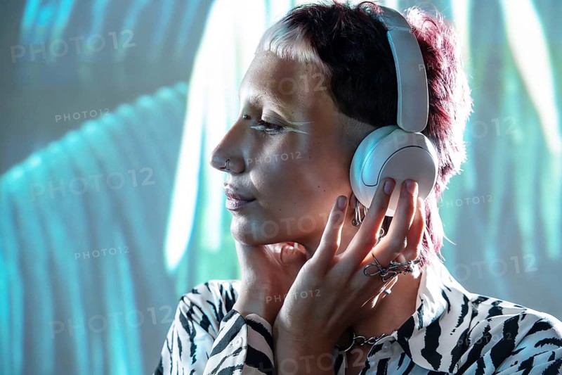 A young, trendy woman with short hair and stylish attire listens to music through headphones, immersed in her own world. The vibrant background complements her modern look