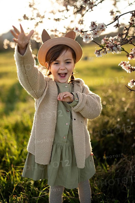 A joyful little girl in a hat is waving and smiling by almond blossoms during a beautiful spring evening, evoking a sense of happiness and the freshness of spring
