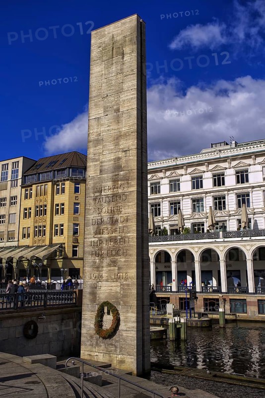 High memorial column with inscription from the First World War on Hamburg's Alster arcades. The war memorial stands right on the water channel, surrounded by historic buildings under blue skies, Hamburg, Germany