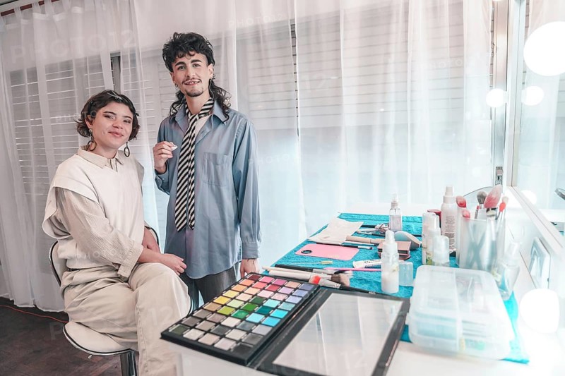 Two makeup artists are getting ready for a photoshoot, surrounded by their tools and equipment in a brightly lit studio, showcasing diversity and inclusion within the beauty industry