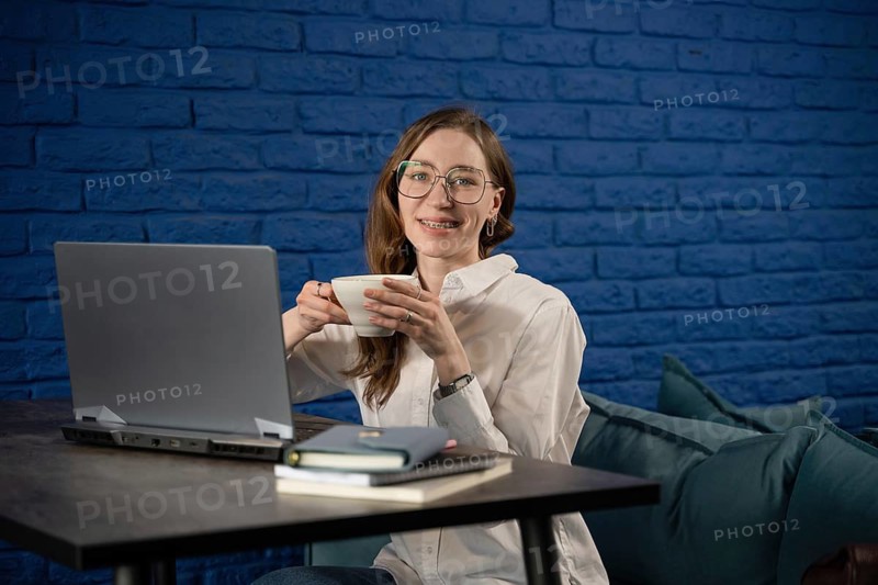 A woman with glasses sits at a desk with a laptop, holding a tea cup, and smiling. The background is a blue brick wall, creating a cozy workspace atmosphere