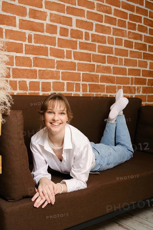 A young woman lies on a brown sofa, smiling. She wears a white shirt and light blue jeans. The background features a brick wall and soft lighting