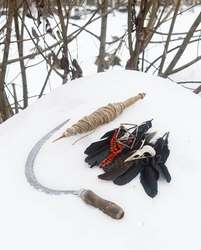 Spindle, crow feathers, bird skull and rowan beads and sickle in the snow. Still life. Pagan, witchcraft symbols