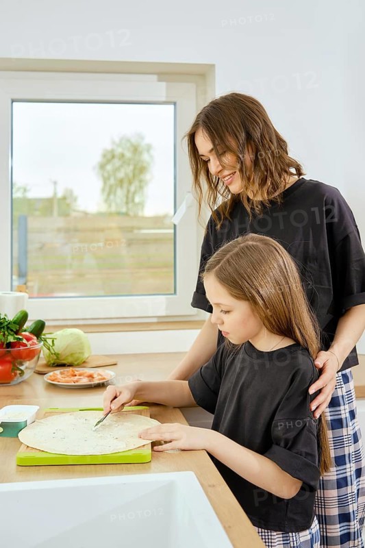 Proud mother watches her young daughter prepare a snack in bright kitchen