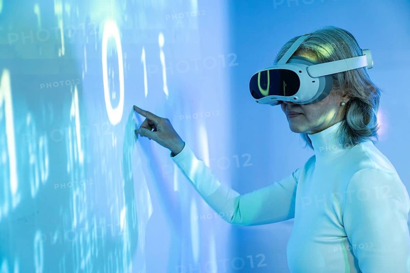 An elderly Asian woman wearing a VR headset interacts with a virtual interface. She navigates the metaverse, demonstrating the fusion of age and advanced technology in digital spaces
