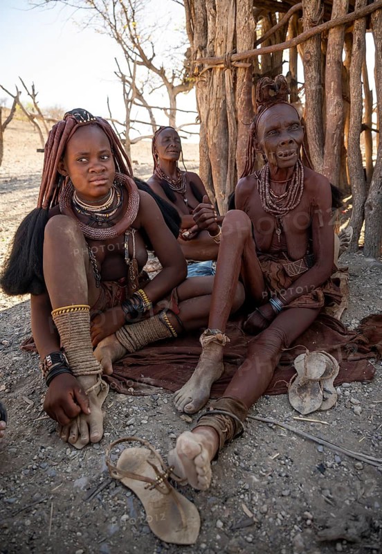 Young and old Himba woman, traditionally dressed, sit in front of a hut in the shade, near Opuwo, Kaokoveld, Kunene, Namibia