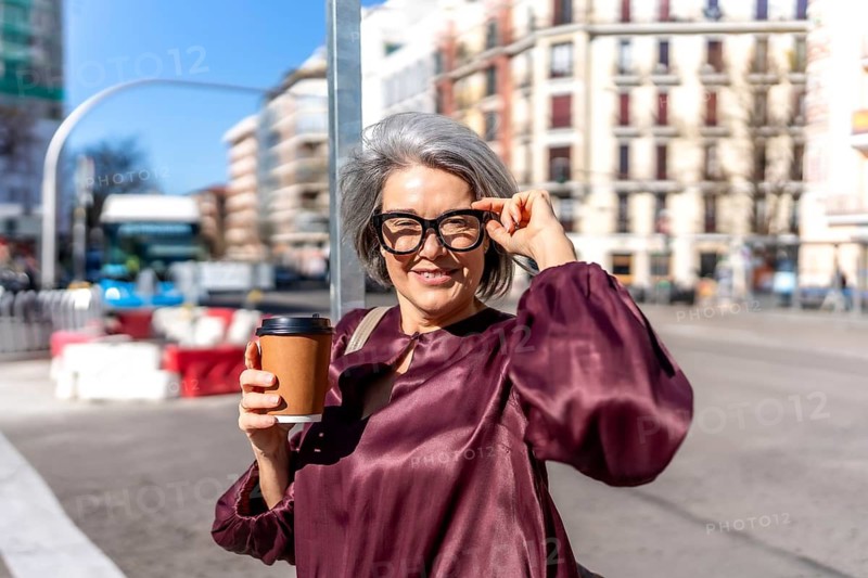 Mature gray haired woman smiling on a busy city sidewalk, adjusting her glasses and holding a takeaway coffee cup while walking through a sunny urban morning bustle