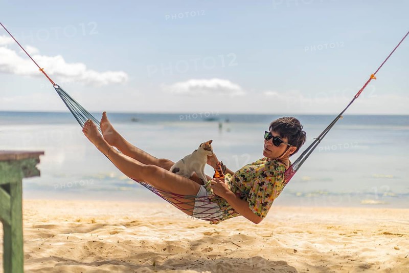 A woman lounges in a colorful hammock on a sandy beach in the Philippines, sharing a playful moment with a small cat under a clear blue sky
