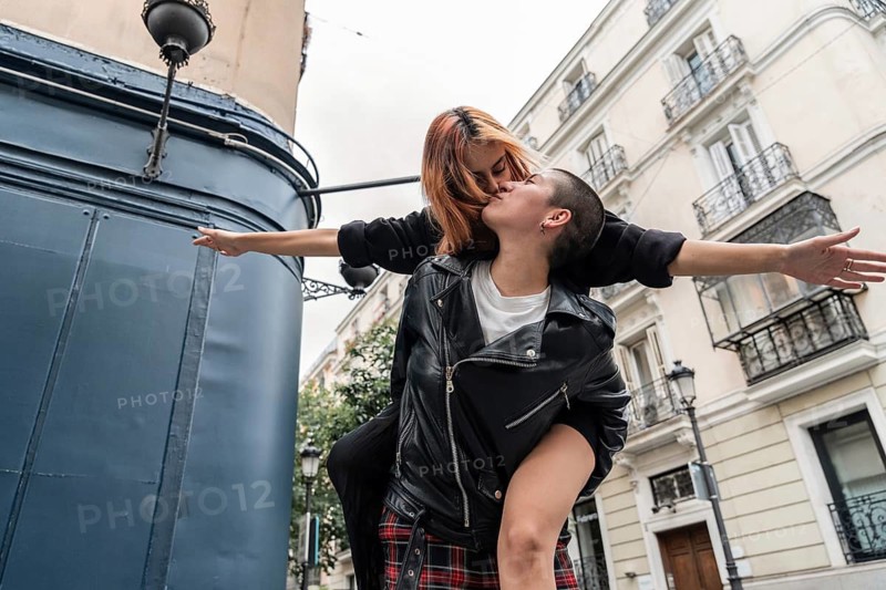 A vibrant couple shares a kiss and playful embrace in a lively urban setting, expressing happiness and love. They're surrounded by picturesque buildings on a cloudy day